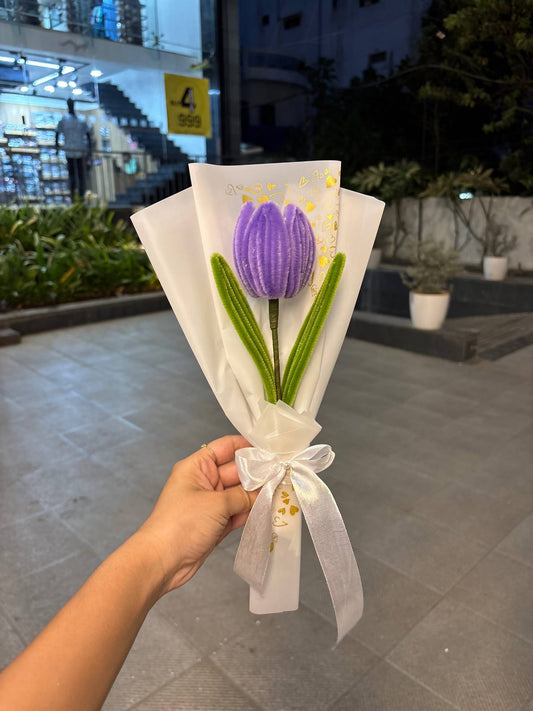 Bouquet of purple tulips wrapped in white paper with a bow, held by a person outdoors.