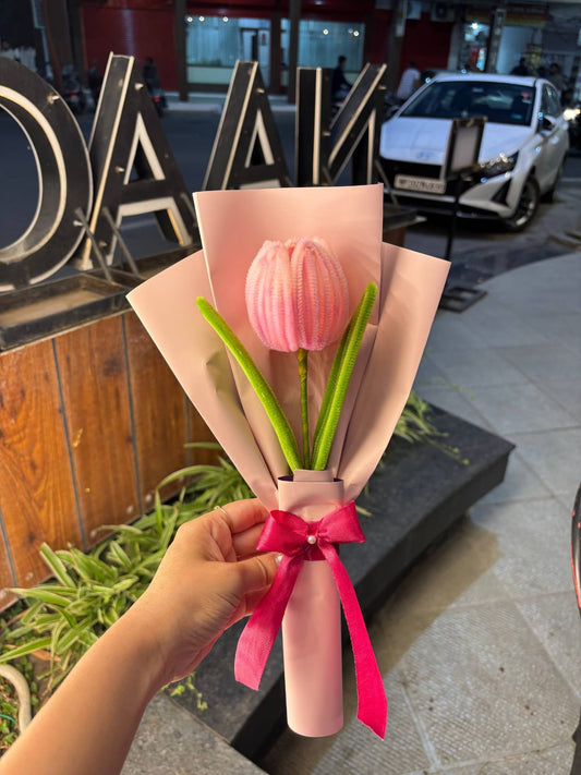 Bouquet of pink tulips wrapped in brown paper with a pink ribbon held by a hand, with a blurred background of a street scene.
