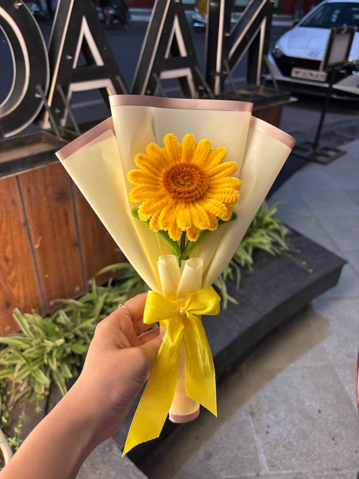 Bouquet of sunflowers wrapped in white paper with a yellow ribbon held by a hand outdoors.
